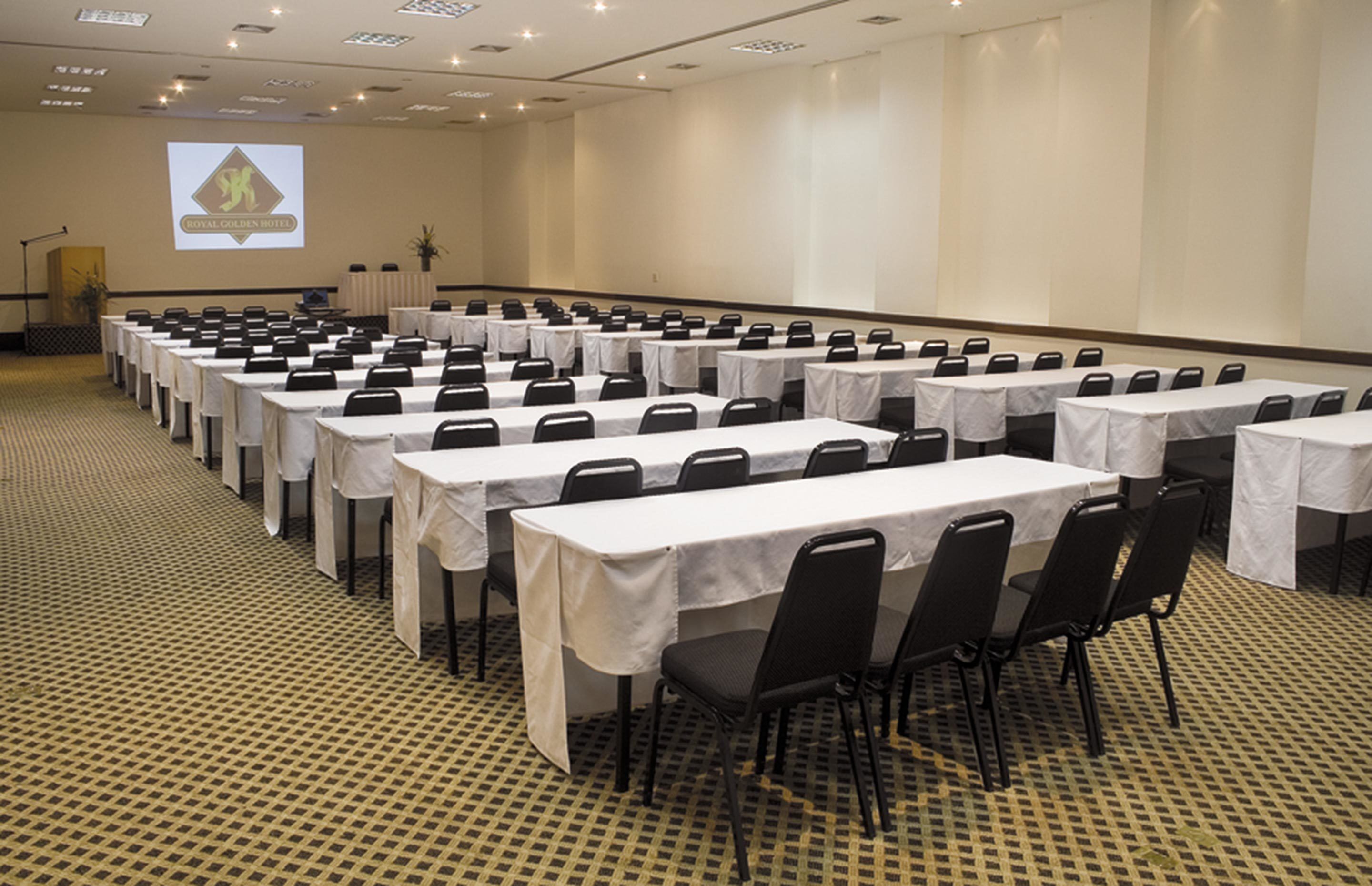 Un salón de conferencias con filas de mesas y sillas, un atril y una pantalla de proyección que muestra el logotipo del Royal Golden Hotel.