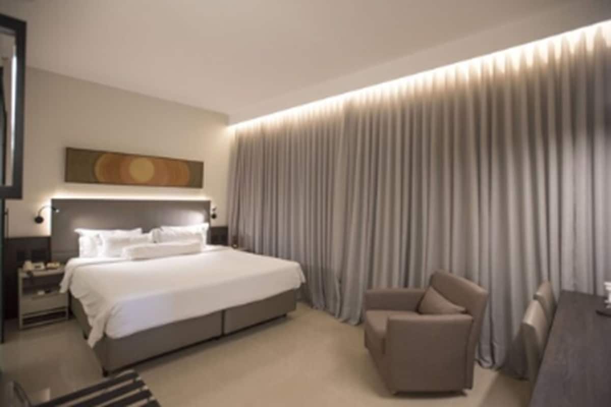 Ein modernes Hotelzimmer mit einem großen Bett, einer Sitzecke und Blick auf die Stadtlandschaft durch das Fenster.