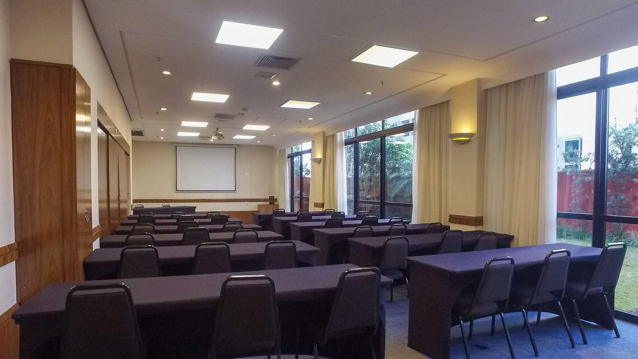A conference room with rows of tables and chairs, a projector screen, and large windows with curtains.