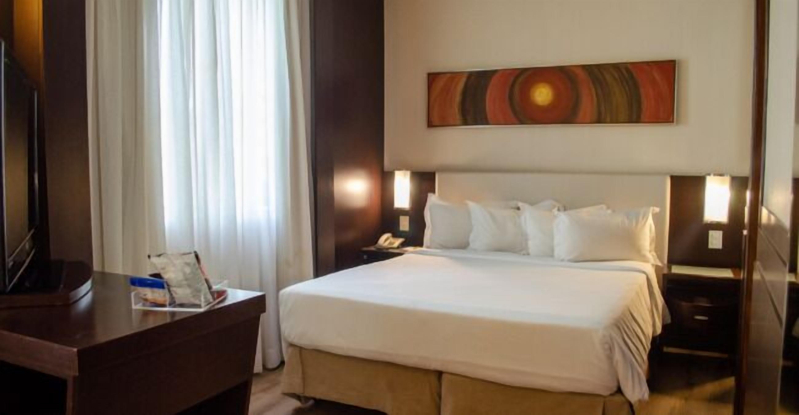 A hotel room with a large bed, a desk with a chair, a telephone, and a painting on the wall.