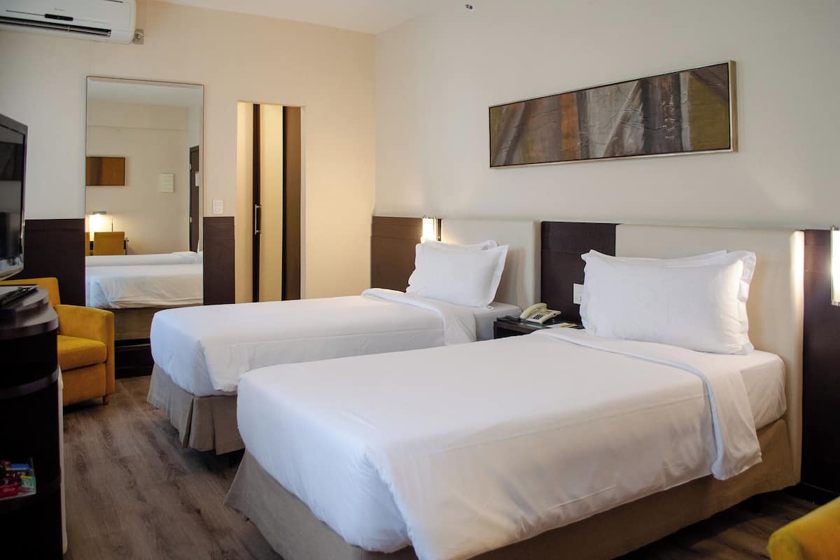 Ein Hotelzimmer mit zwei Einzelbetten, einem Schreibtisch mit Stuhl, einem Fernseher und einem Bild an der Wand.