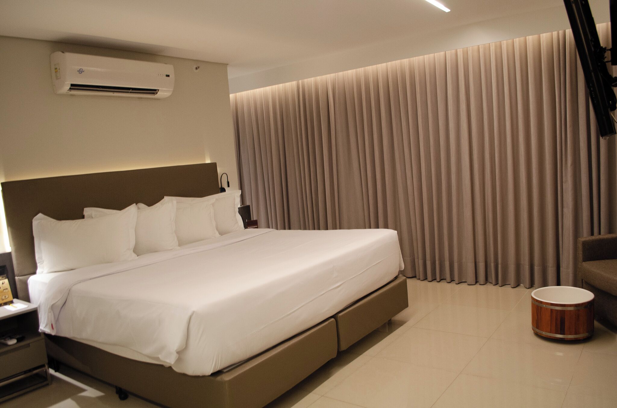 A modern hotel room with a large bed, an air conditioning unit, a bedside table, a lamp, and a drum.