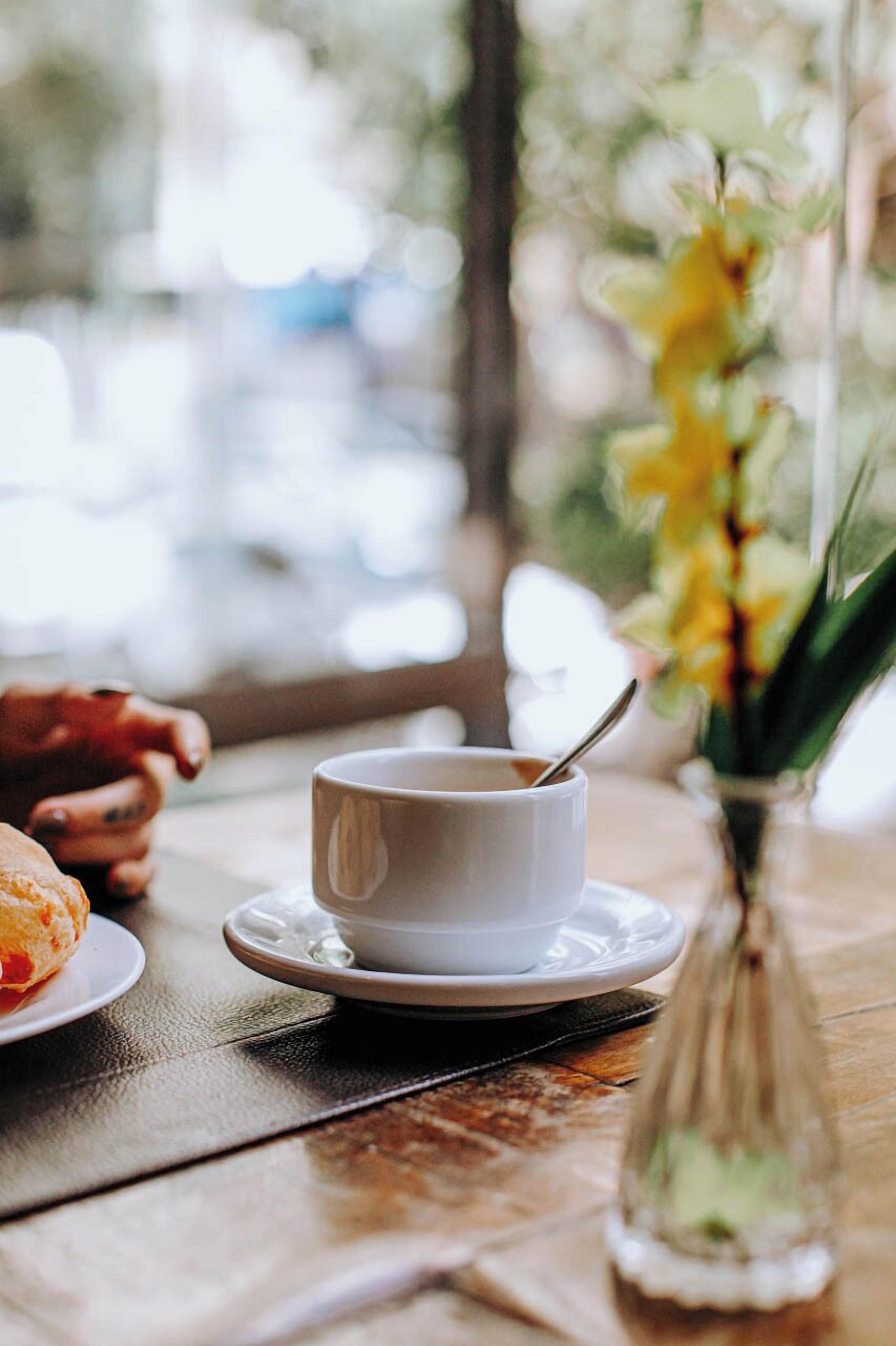 Uma mesa com uma xícara de café, um prato de comida e um vaso de flores.