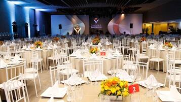 A banquet hall with round tables, white chairs, and floral centerpieces.