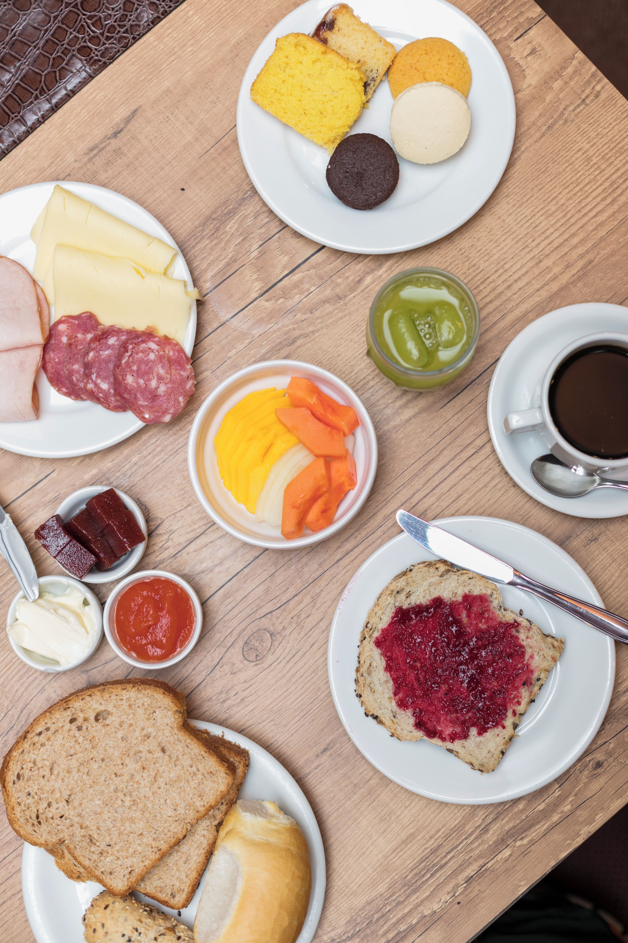 Uma mesa de madeira com diversos itens de pequeno-almoço, incluindo pão, queijo, carnes e uma chávena de café.