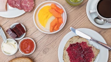 Uma mesa de madeira com diversos itens de pequeno-almoço, incluindo pão, queijo, carnes e uma chávena de café.