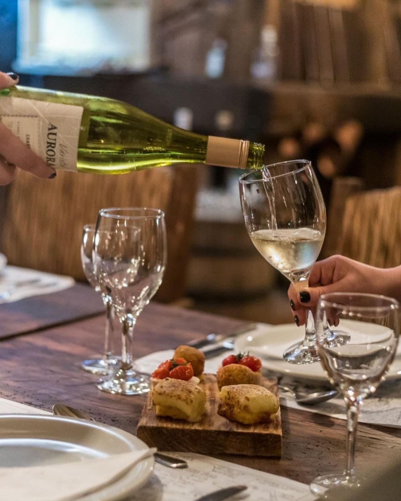 A person pouring wine into a glass. There are empty glasses and a wooden board with food items on the table.