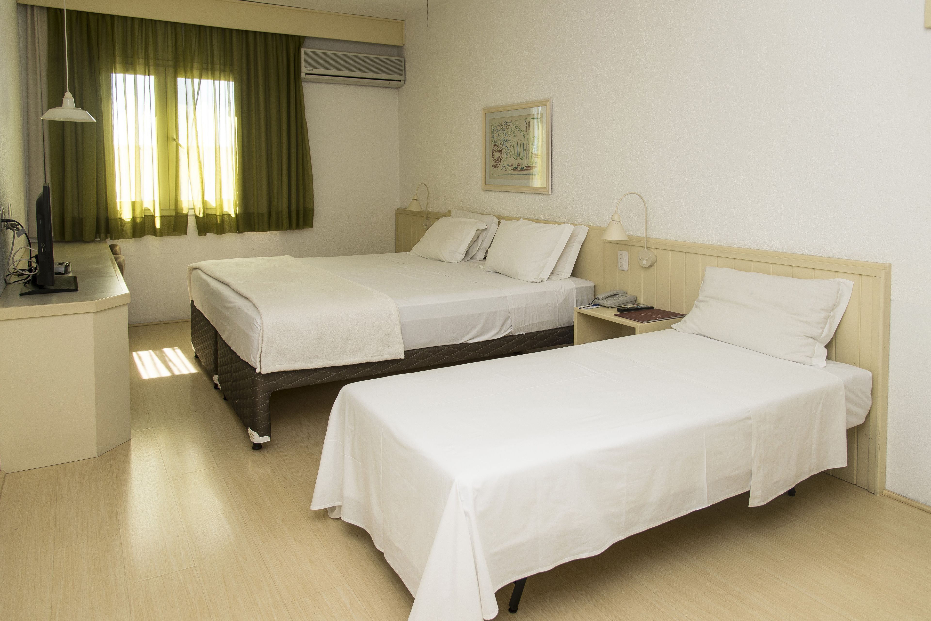 Quarto de hotel com duas camas de solteiro, uma escrivaninha com computador, abajur e janela com cortinas.