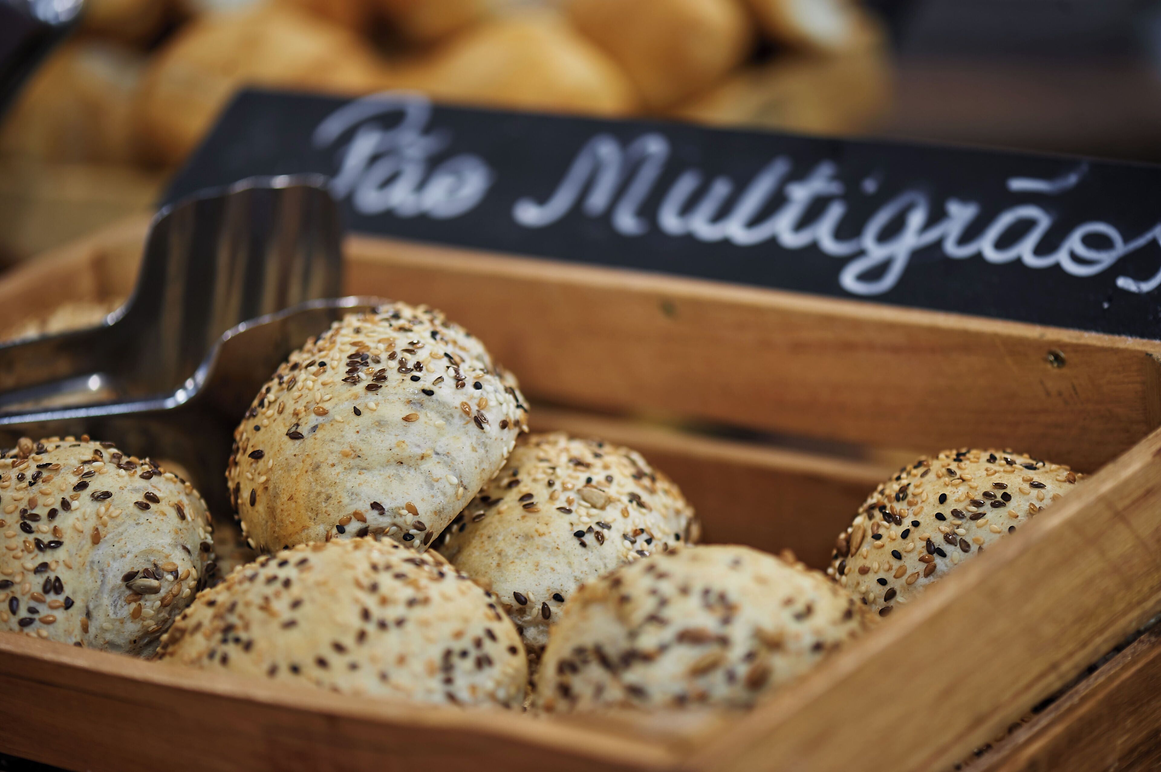 A wooden box filled with round bread rolls topped with seeds, with a sign reading "Pão Multigraos" in the background.