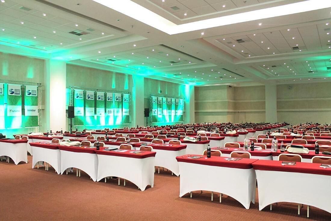 Un amplio salón de conferencias con filas de mesas y sillas, iluminación ambiental verde y un escenario con un atril.