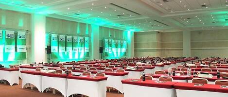 Un amplio salón de conferencias con filas de mesas y sillas, iluminación ambiental verde y un escenario con un atril.