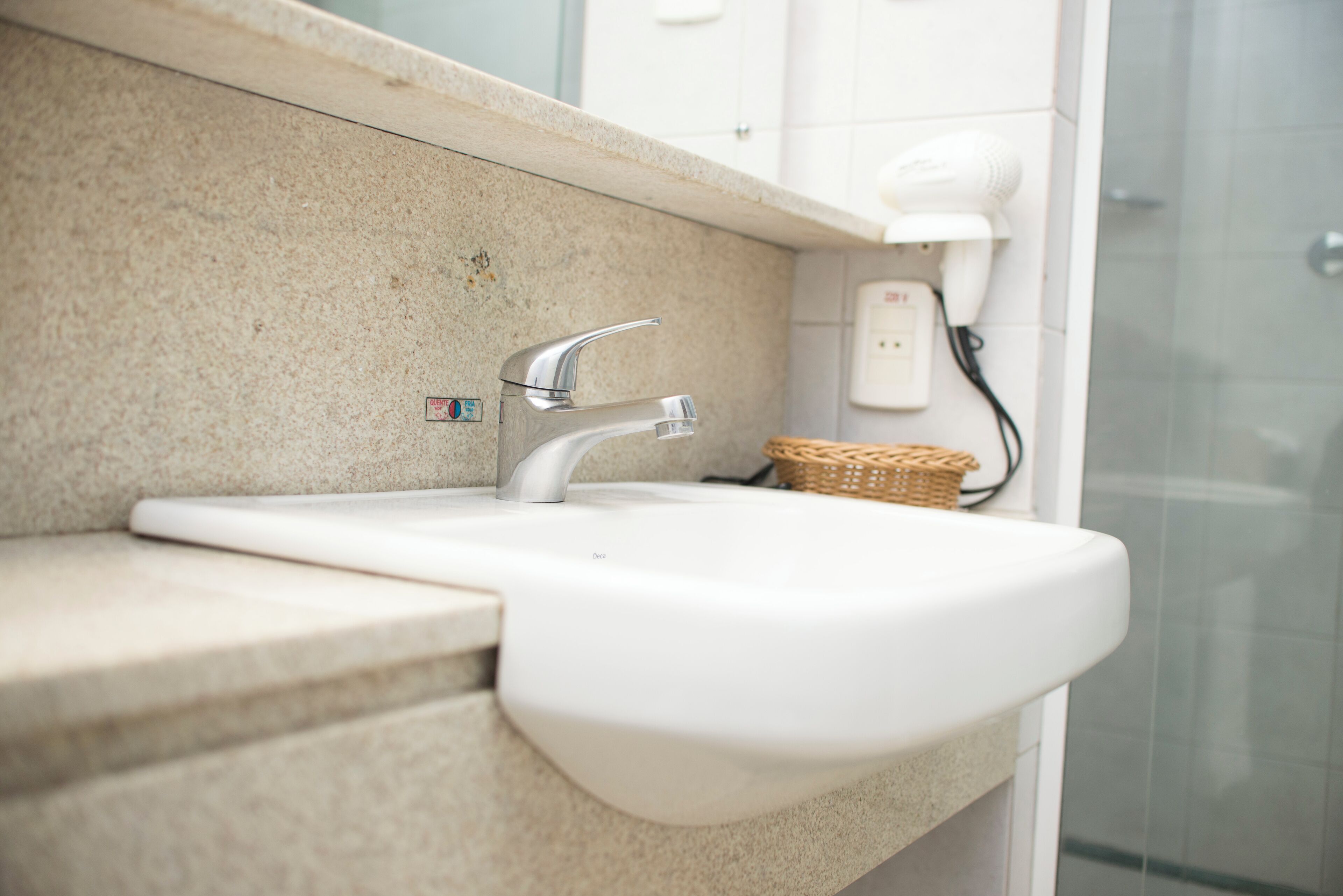 A bathroom with a white sink, a chrome faucet, a wicker basket, and a hairdryer on the wall.