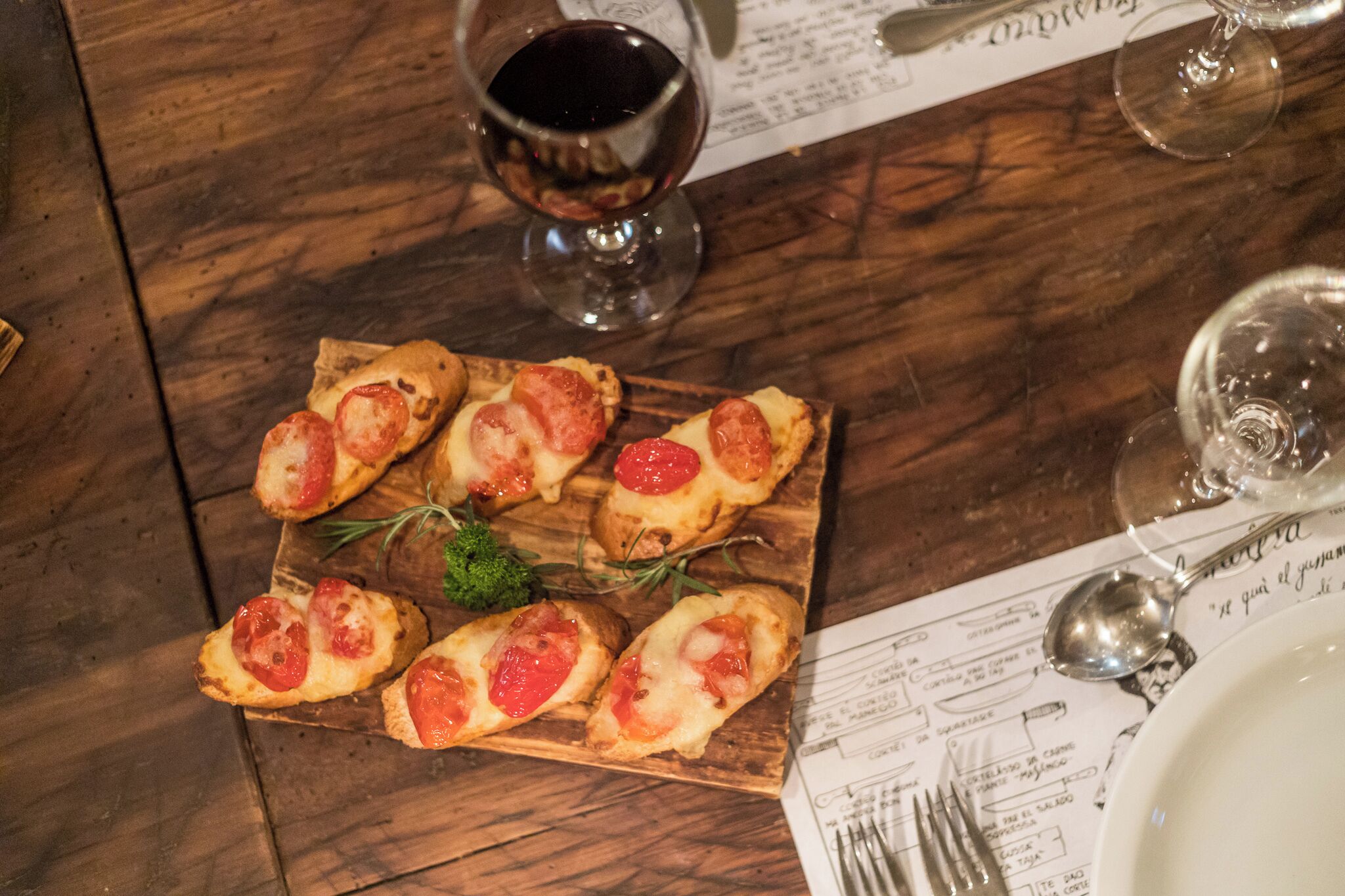 Una tabla de madera con varios bocadillos de bruschetta cubiertos de tomates y queso, una copa de vino tinto, y un menú sobre una mesa de madera.