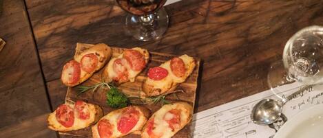 Una tabla de madera con varios bocadillos de bruschetta cubiertos de tomates y queso, una copa de vino tinto, y un menú sobre una mesa de madera.