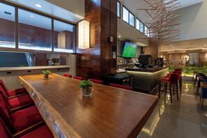 Zona de bar moderna con una mesa de madera larga, asientos acolchados de color rojo y una pantalla de televisión mostrando un partido deportivo.