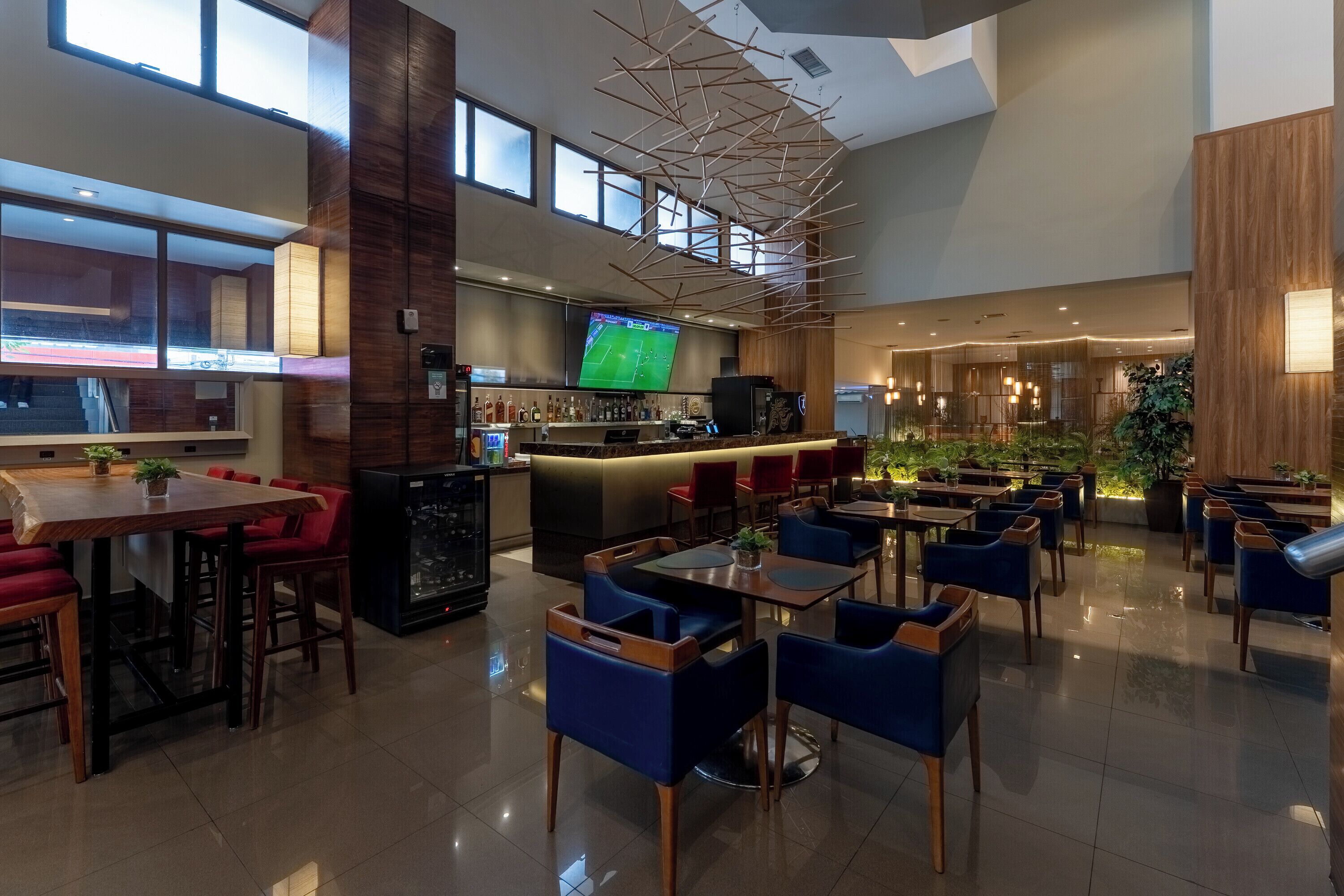 A modern hotel lobby with a bar area, seating arrangements, and a large screen displaying a sports event.
