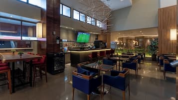 A modern hotel lobby with a bar area, seating arrangements, and a large screen displaying a sports event.