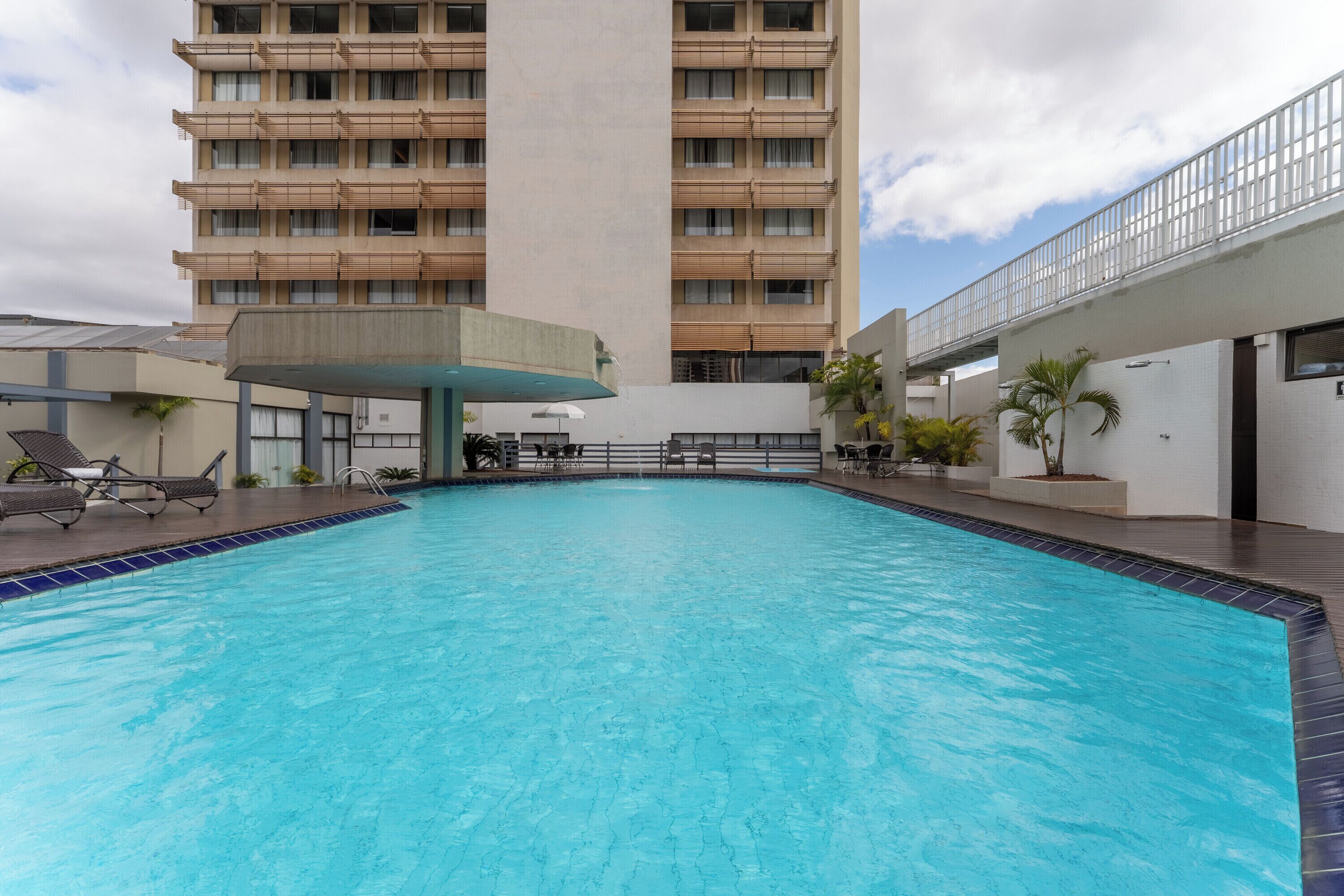 Piscina com espreguiçadeiras, área de estar coberta e um edifício alto ao fundo.