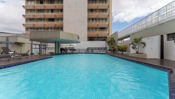 Piscina com espreguiçadeiras, área de estar coberta e um edifício alto ao fundo.