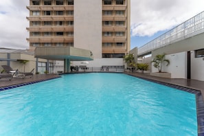 Piscina con sillas de descanso, zona de estar techada y un edificio alto al fondo.