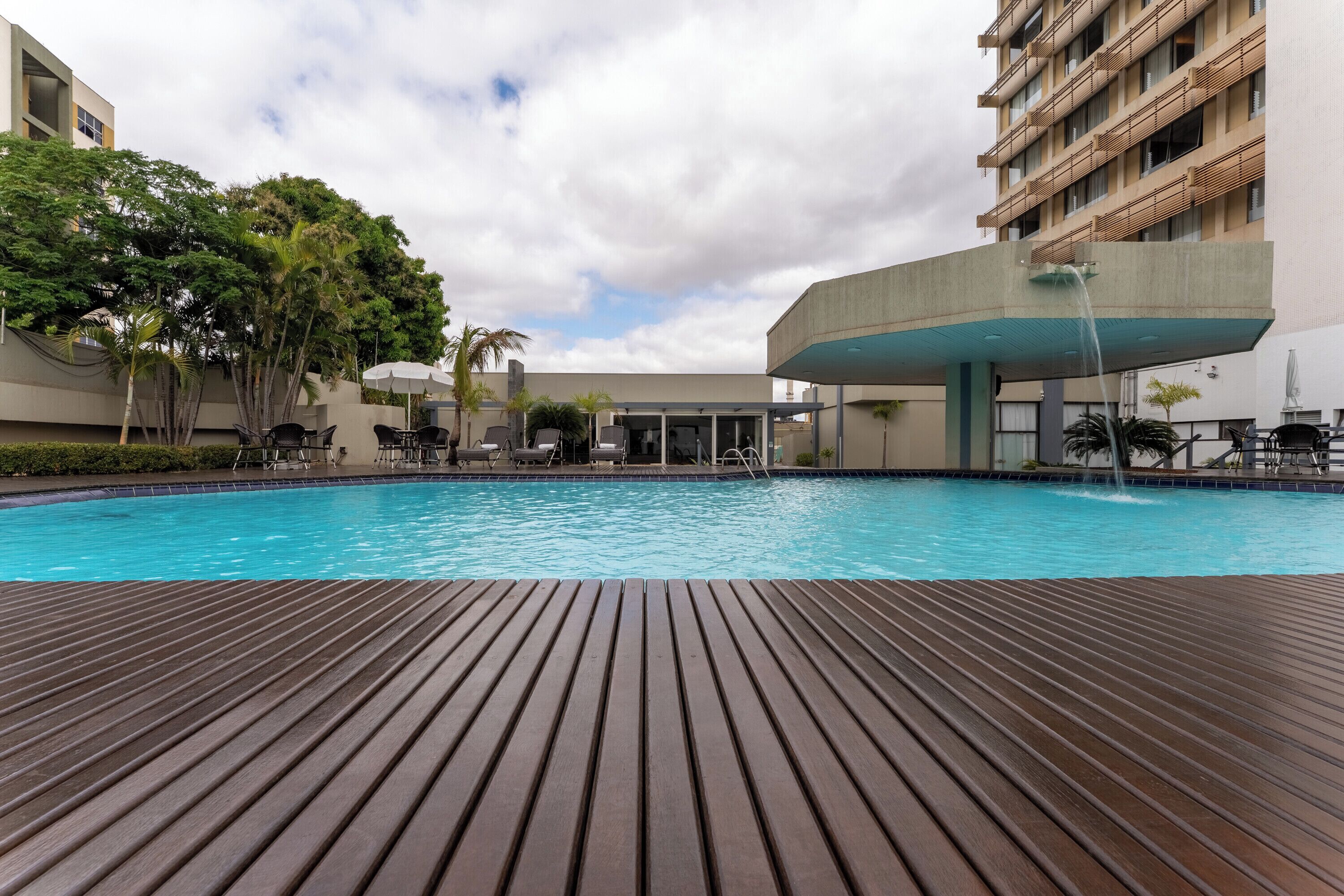 Una piscina con cubierta de madera, rodeada de sillas de descanso y sombrillas, con edificios al fondo.