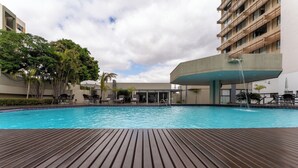 A swimming pool with a wooden deck, surrounded by lounge chairs and umbrellas, with buildings in the background. - Hotel Deville Prime Cuiabá (Cuiabá)
