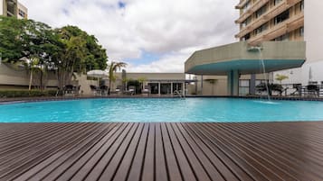 Una piscina con cubierta de madera, rodeada de sillas de descanso y sombrillas, con edificios al fondo.