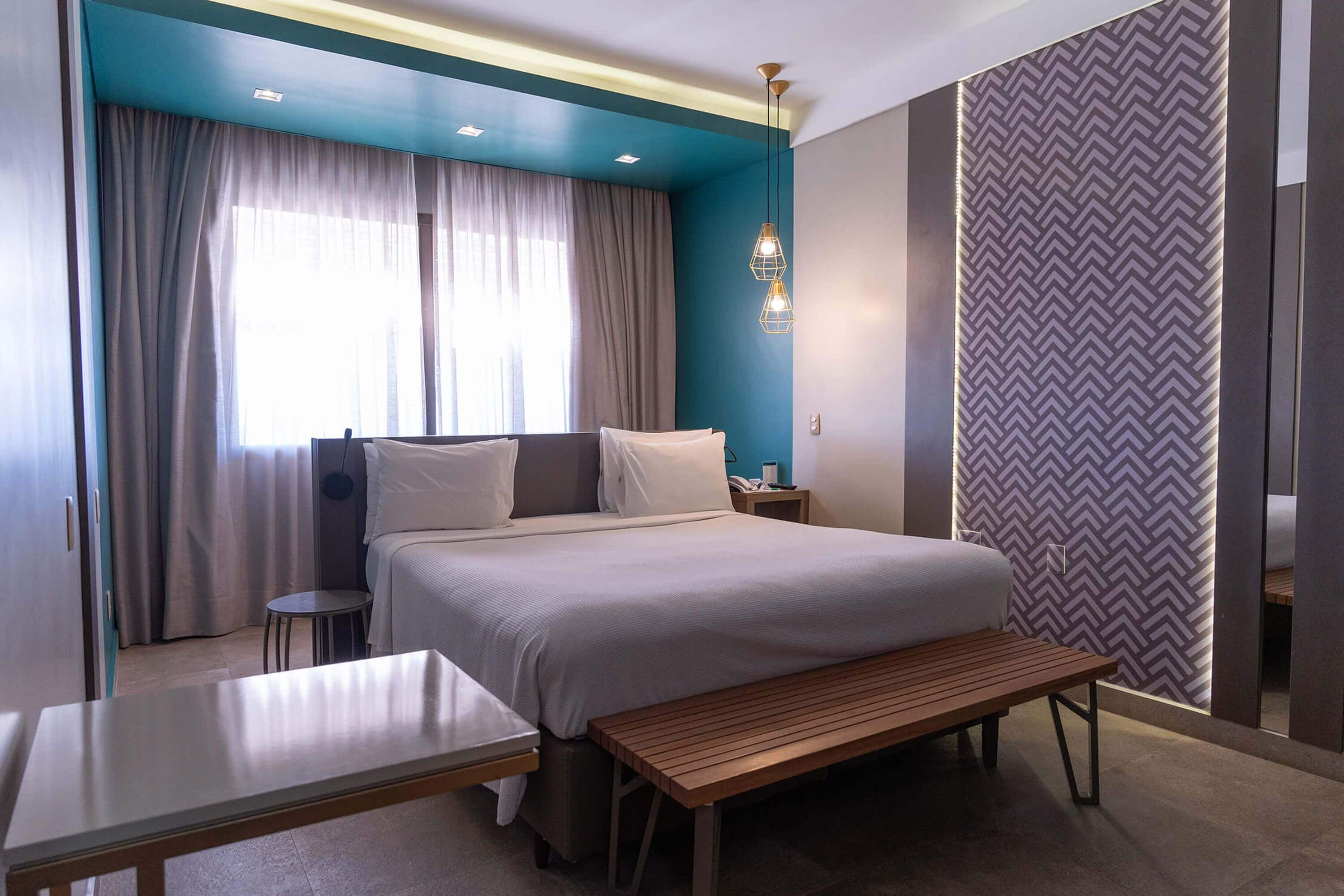 Habitación de hotel moderna con cama, mesita de noche, banco y cortinas.