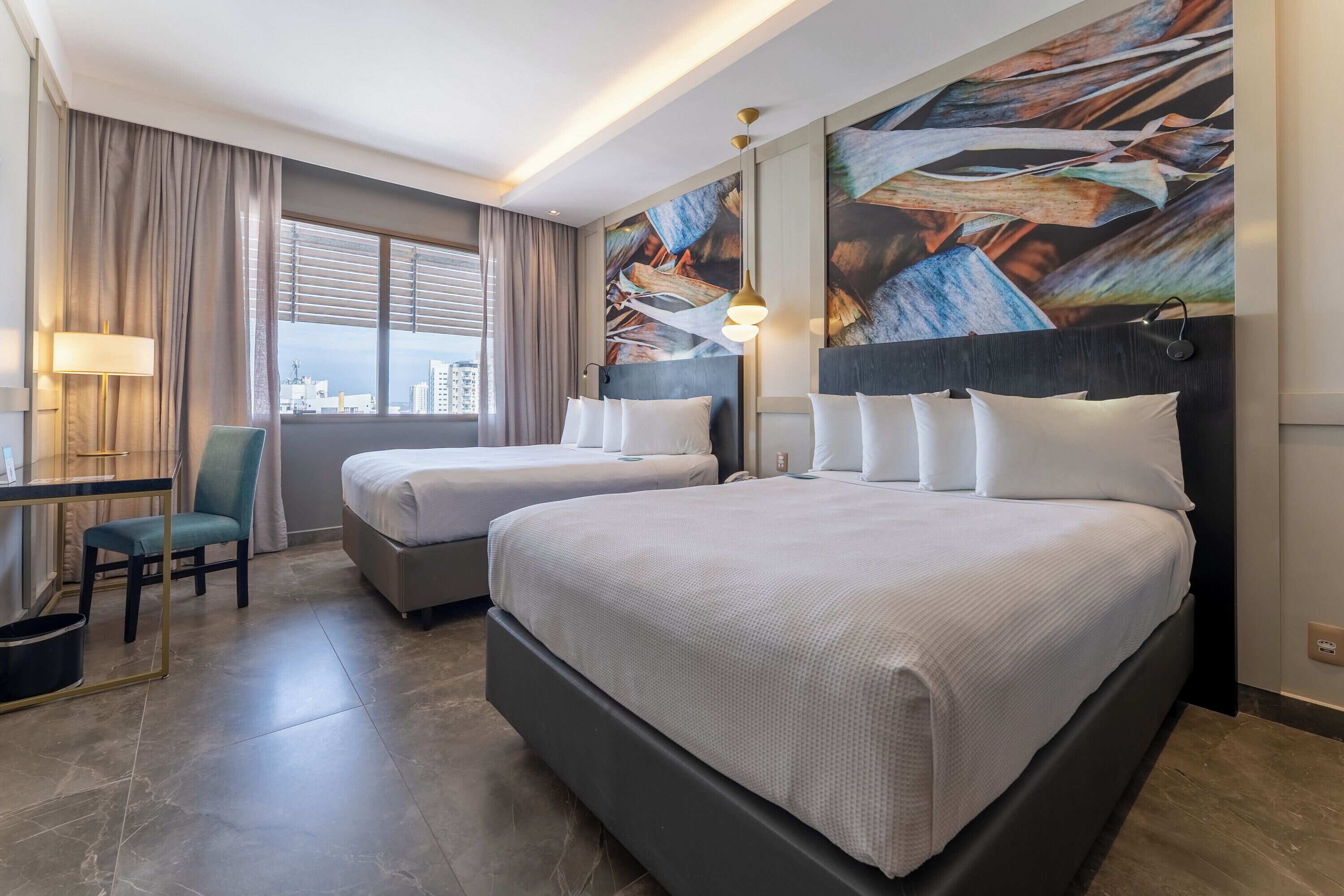 Una camera d'albergo con due letti, una scrivania, una sedia e un'ampia finestra con tende.