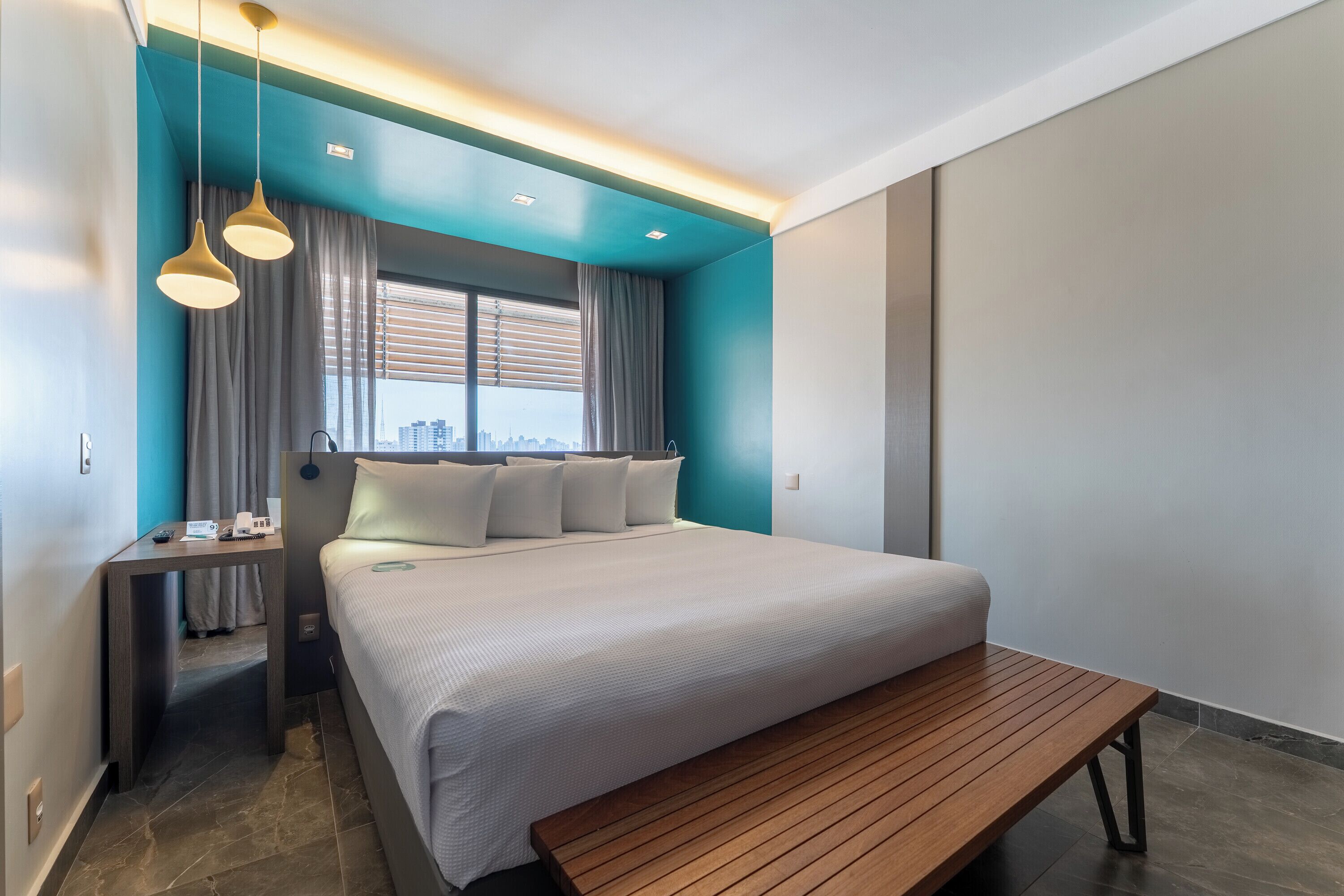 Habitació d'hotel moderna amb un llit gran, una banca de fusta, un escriptori amb un telèfon i vistes a la ciutat.