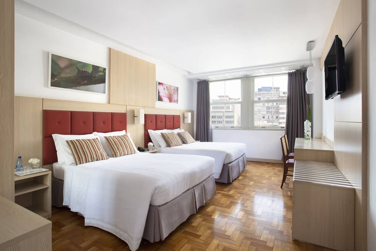 A hotel room with two beds, a TV, a window with a view of buildings, and a wooden floor.