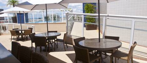 An outdoor patio with round tables and wicker chairs, umbrellas, and a view of a pool and buildings.
