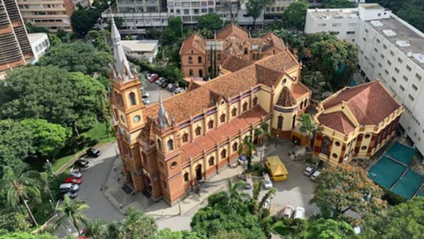 Un edifici gran i històric amb una torre destacada i diverses ales, envoltat de vegetació exuberant i edificis moderns.