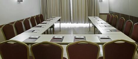 Un salón de conferencias con una larga mesa y sillas, ventanas amplias con cortinas e iluminación en la pared.