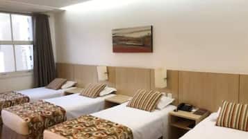 A hotel room with three beds, each with a patterned bedspread and matching pillows. There is a painting on the wall above the beds, a window with curtains, and a nightstand with a phone and a book.
