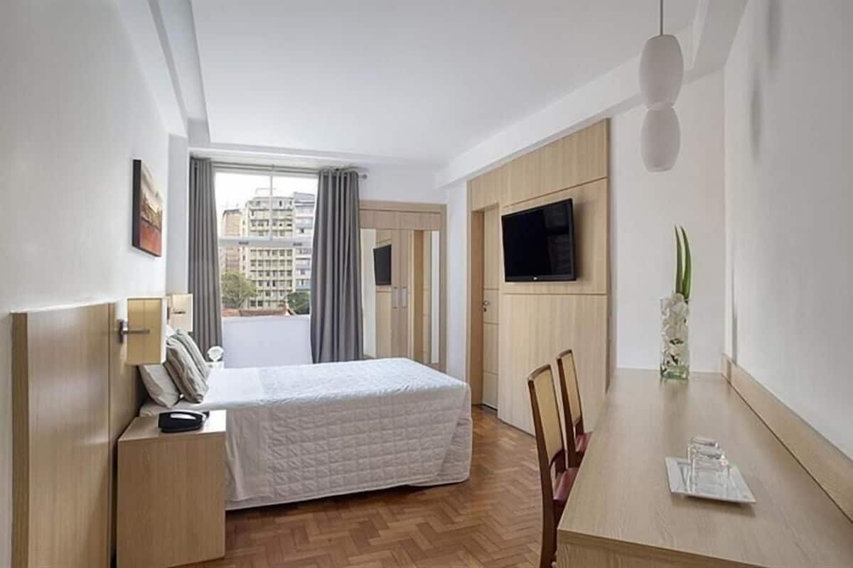 A hotel room with a bed, a desk, a chair, a TV, and a window with curtains.