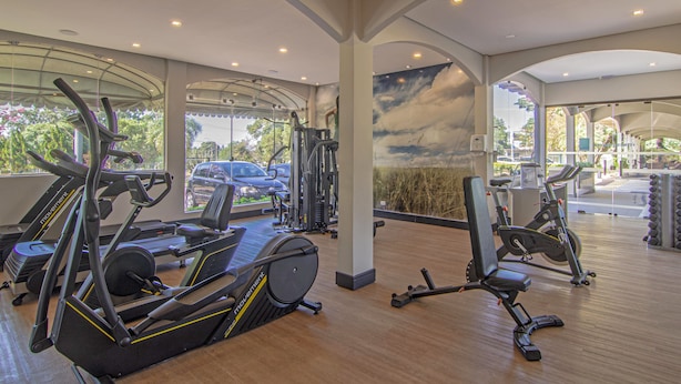Un gimnasio espacioso con diversas máquinas de cardio, un mural en la pared y grandes ventanales que ofrecen vistas al exterior.