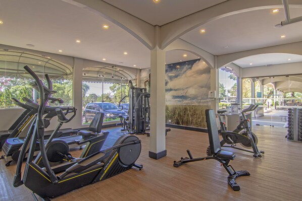 A spacious gym with various cardio machines, a wall mural, and large windows offering views of the outside. - Hotel Deville Express Cascavel (Cascavel)