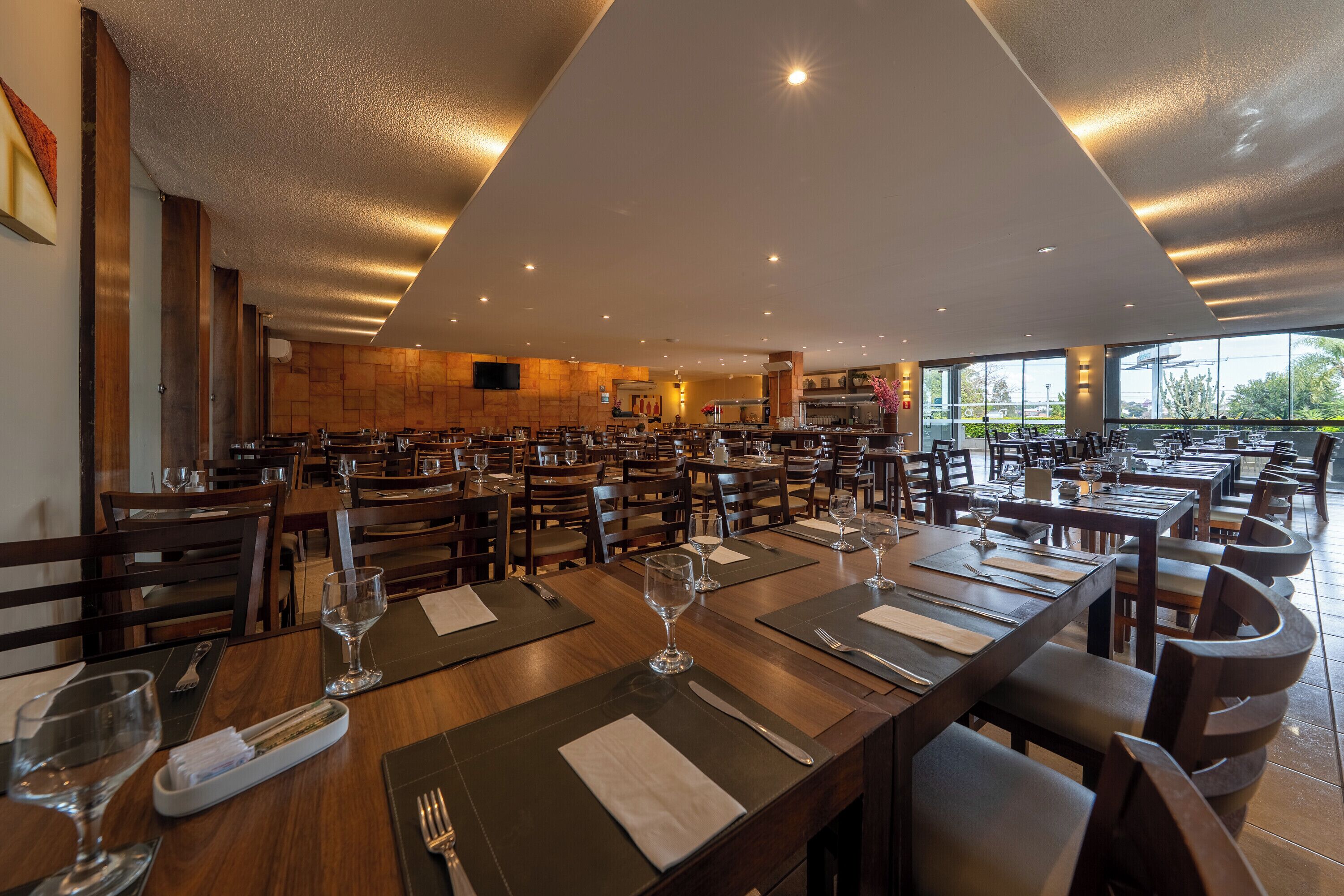 A spacious restaurant with multiple dining tables and chairs, a modern ceiling design, and large windows offering a view of the outdoors.