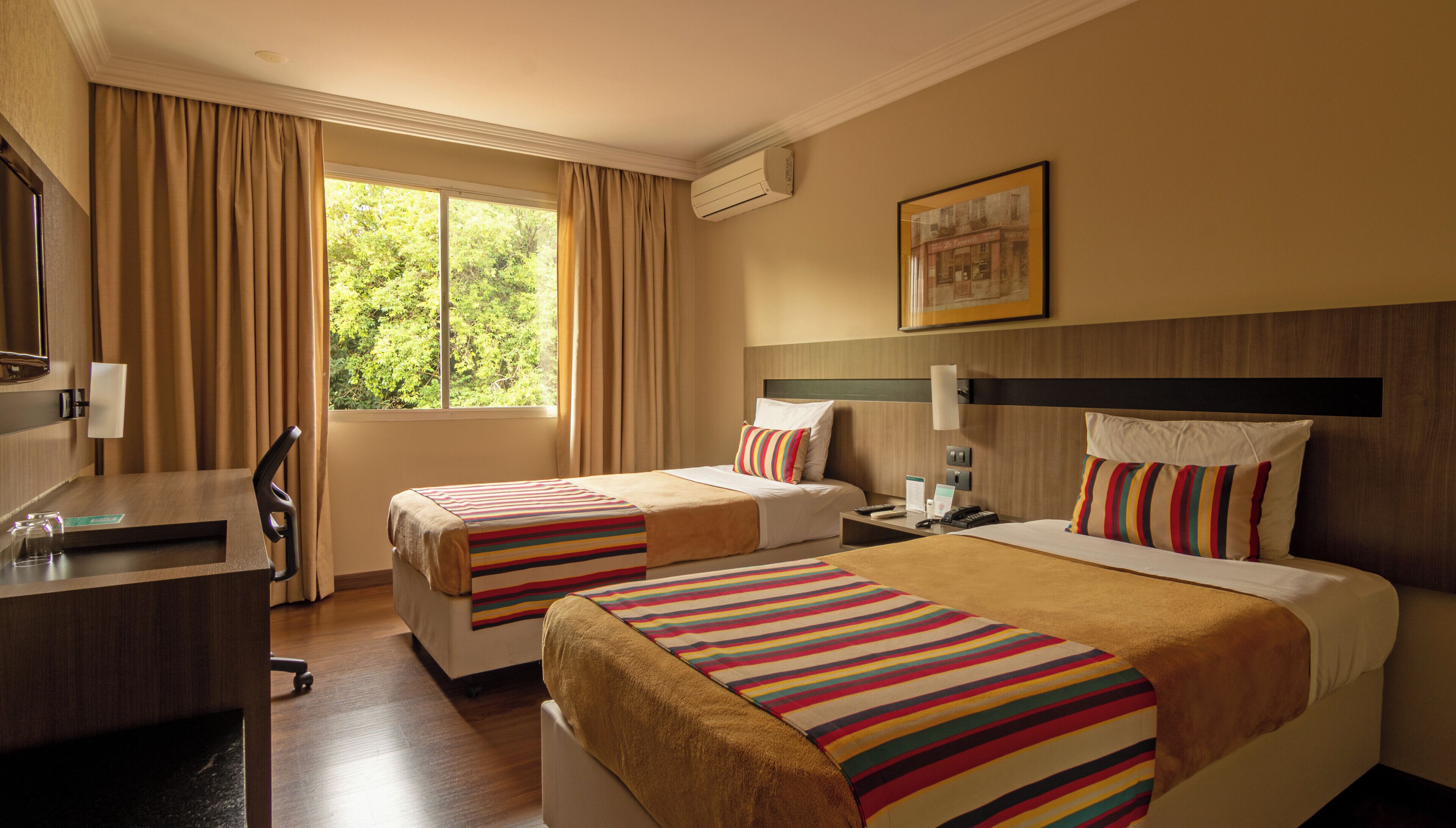 Habitación de hotel con dos camas, un escritorio, una silla, una ventana con cortinas, un equipo de aire acondicionado y un cuadro en la pared.