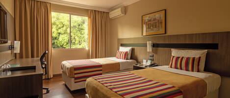 Habitación de hotel con dos camas, un escritorio, una silla, una ventana con cortinas, un equipo de aire acondicionado y un cuadro en la pared.