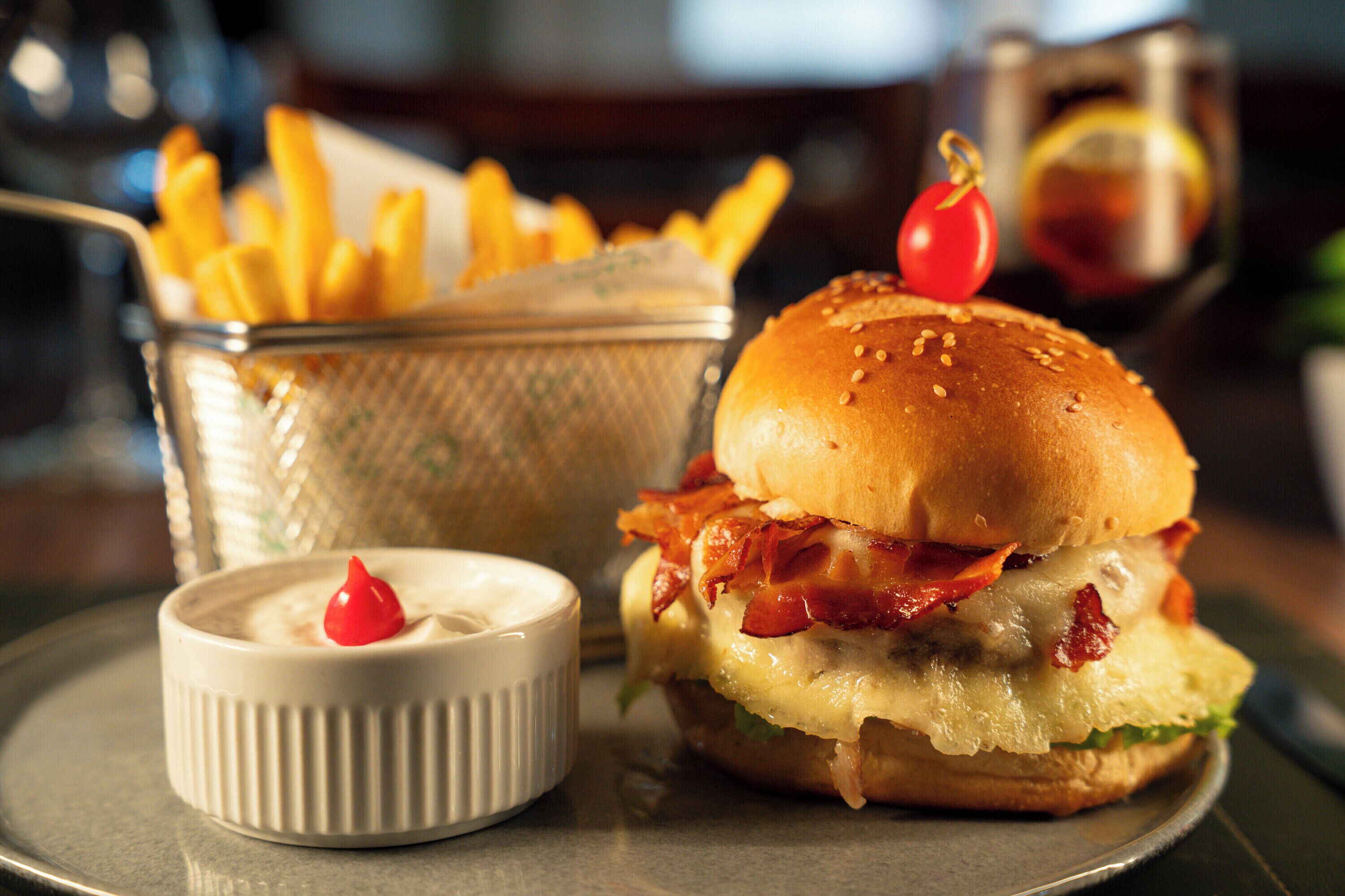 Hamburguesa con tocino, queso y tomate, acompañada de papas fritas y una salsera pequeña.