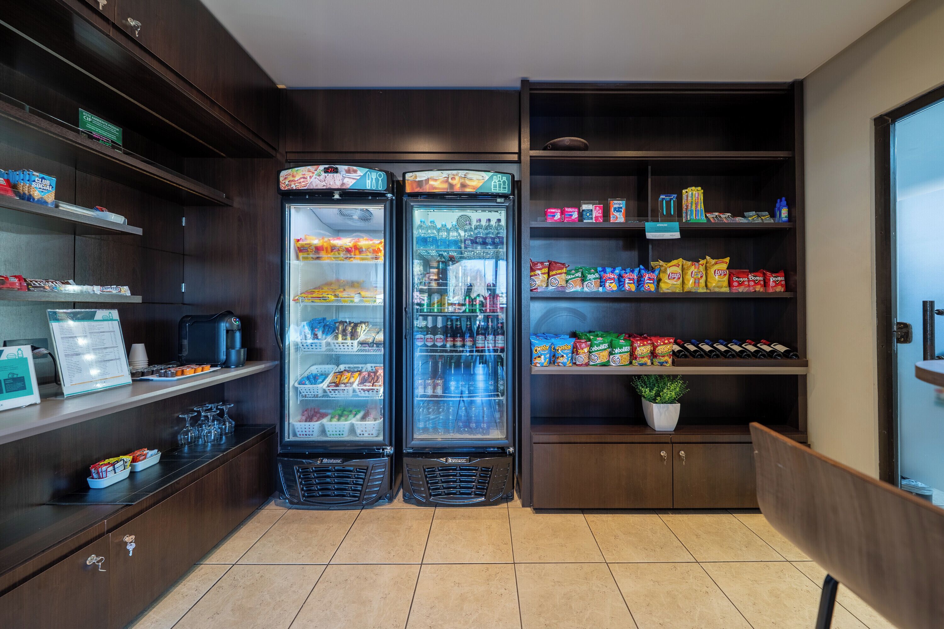 Una despensa de hotel con dos refrigeradores llenos de refrigerios, una máquina de café y una planta en maceta.