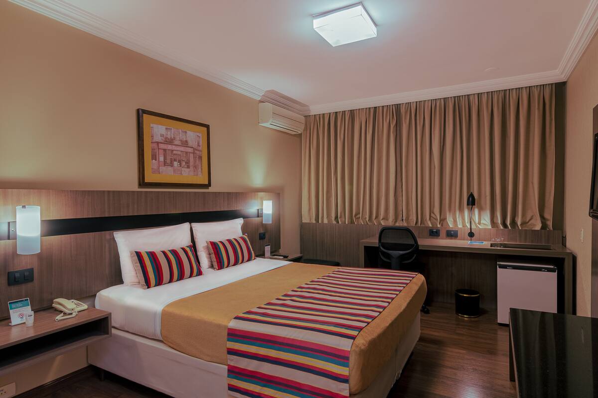 A hotel room with a bed, a desk, a chair, a small refrigerator, and a trash can.