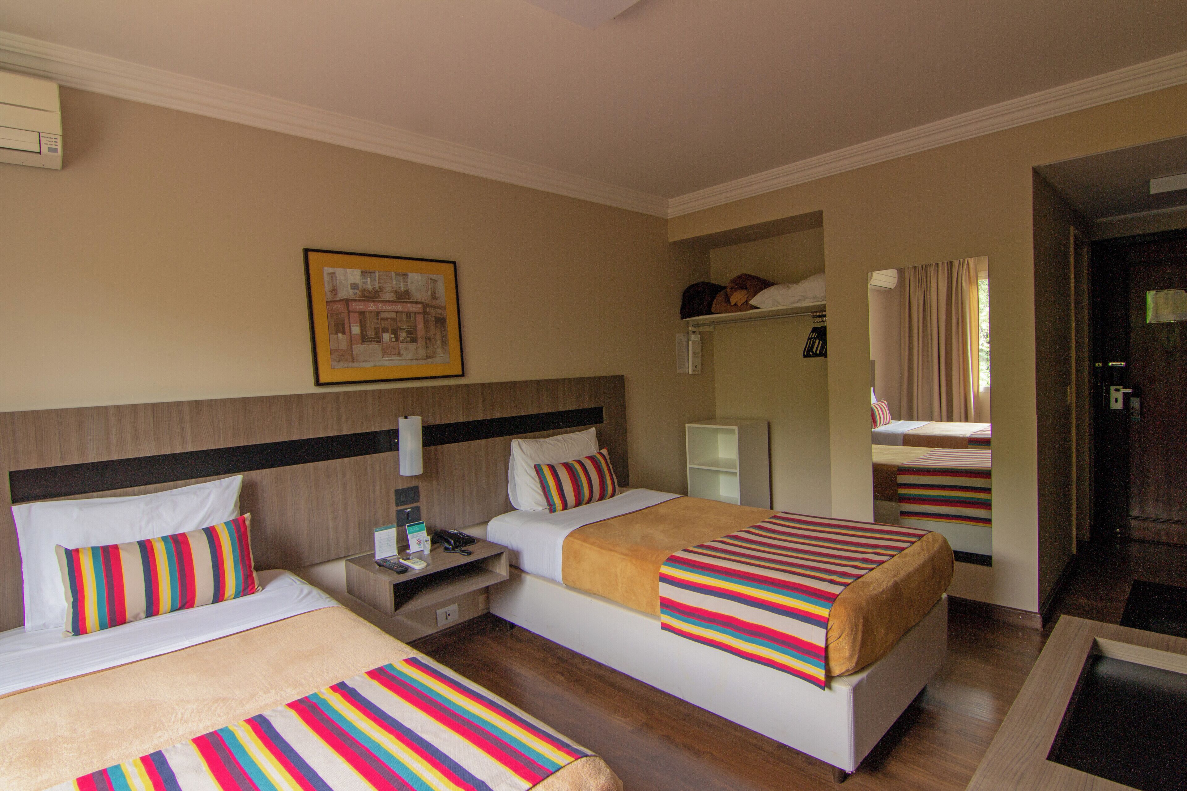 Habitación de hotel con dos camas, una mesita de noche, una lámpara, un cuadro enmarcado en la pared y una ventana con cortinas.