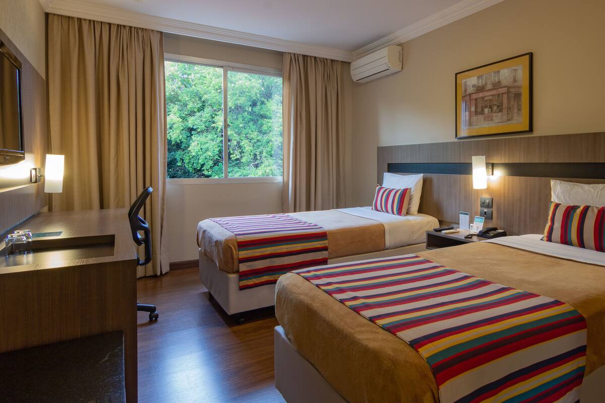 A hotel room with two beds, a desk, a chair, a lamp, and a window with curtains.