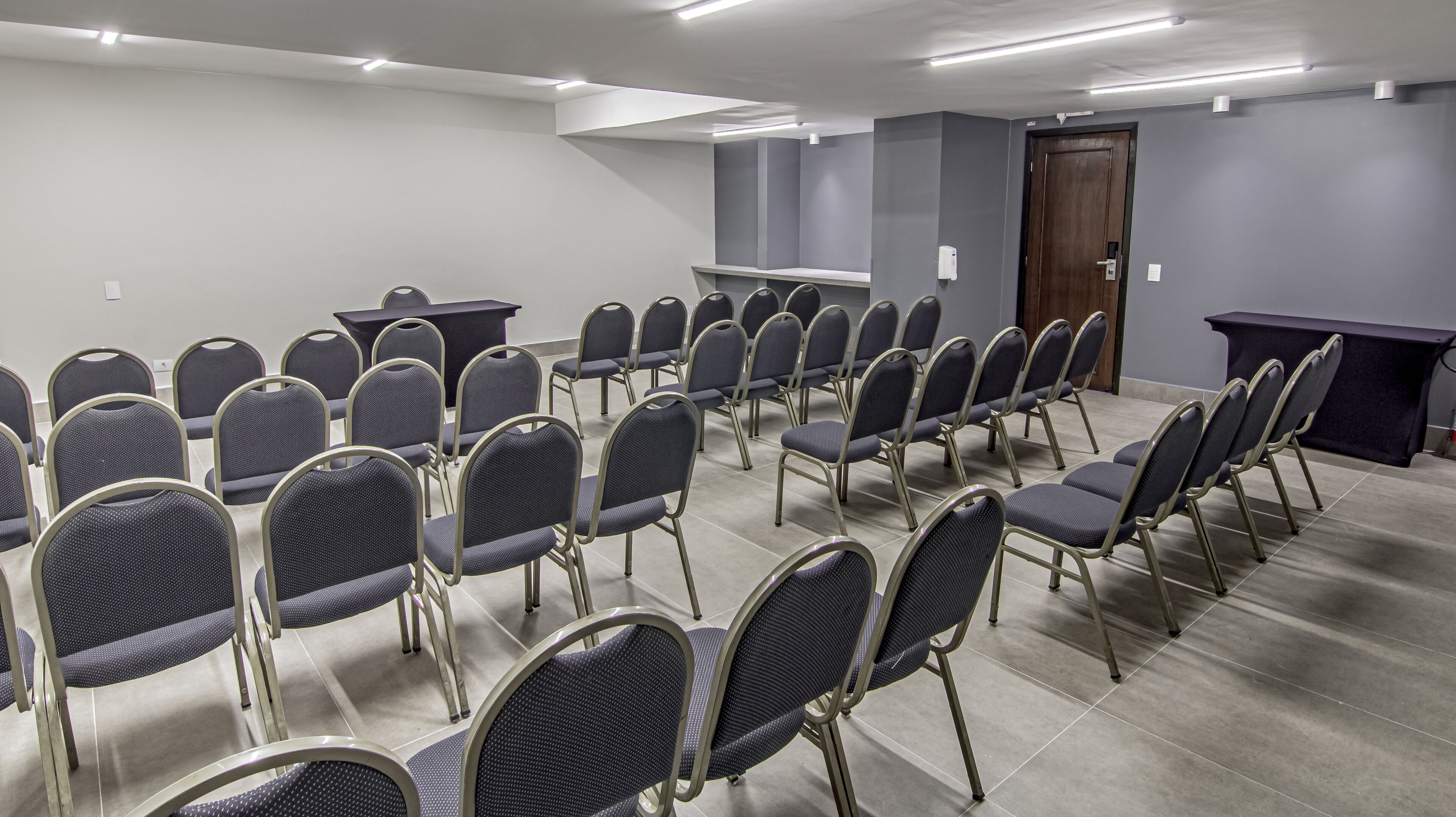 A conference room with rows of chairs and a table, grey walls, and a door.