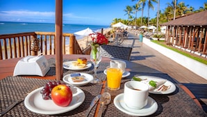 Una mesa de desayuno con platos de comida, un vaso de jugo de naranja y una taza blanca, con vista a una playa con palmeras y un cielo despejado.