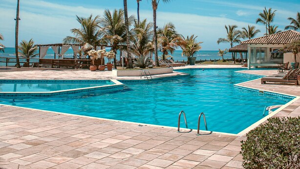 Zona de piscina con agua cristalina azul, rodeada de palmeras y vista al océano.