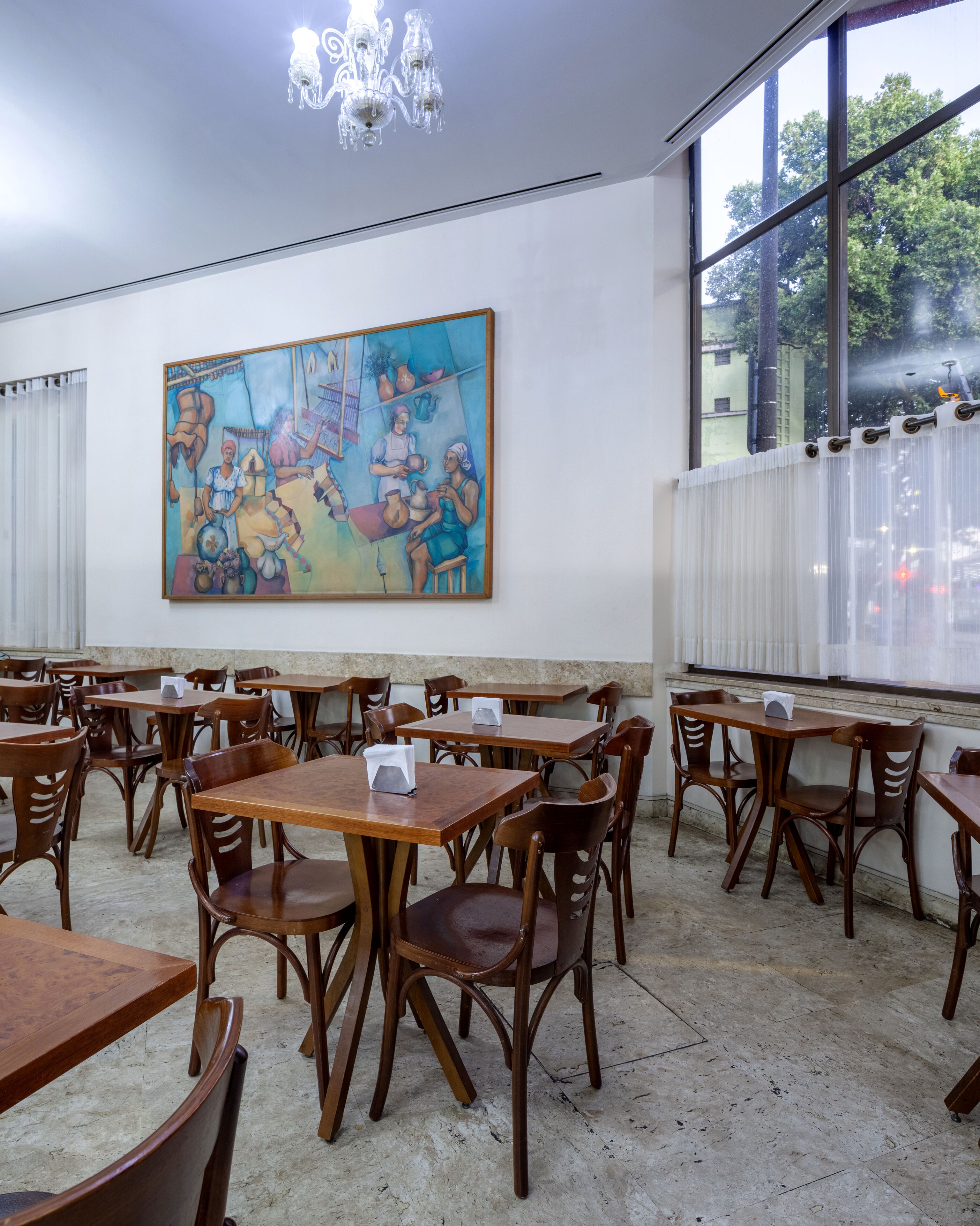 A dining area with wooden tables and chairs, a large painting on the wall, and a view through the window.